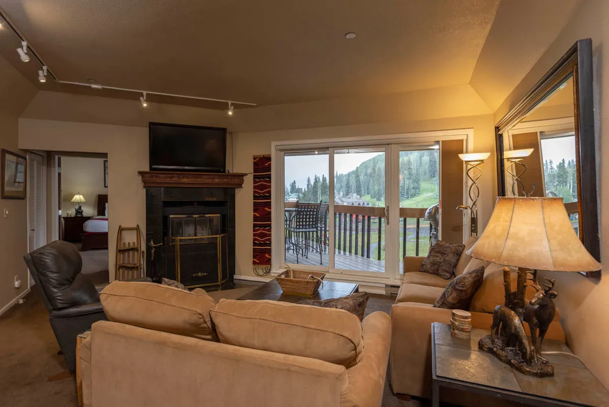 This image showcases a spacious living area featuring a stone fireplace, large windows that let in abundant natural light, and a comfortable seating arrangement perfect for gatherings. The view through the windows reveals majestic mountain scenery, directly tying into the room's ambiance and the outside environment.