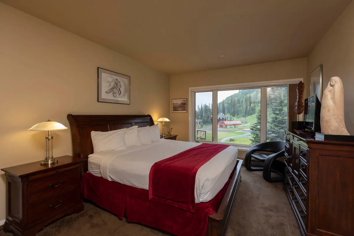 This cozy bedroom features a queen-sized bed, dark wooden furniture, and a large window offering sweeping mountain vistas. The inviting arrangement is perfect for relaxing after a day on the slopes or enjoying a peaceful start to the morning.
