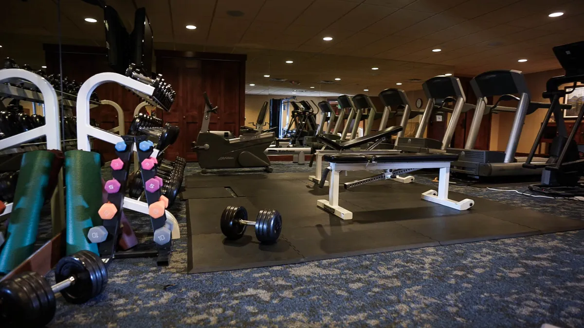 An alternate view of the fitness center, focusing on dumbbells, treadmills, a weight bench, and exercise mats arranged for both solo and group activities. The space is designed to motivate and foster community interaction.