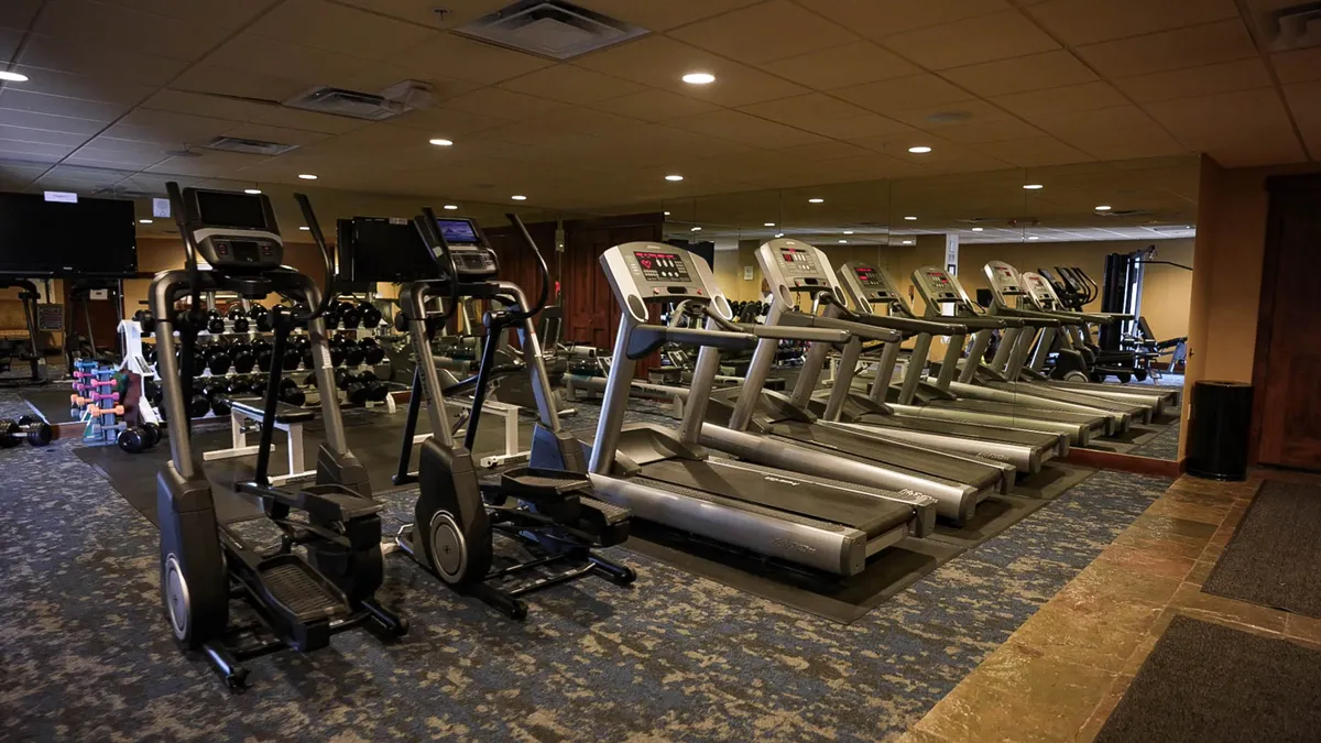 A modern fitness center interior featuring treadmills, elliptical machines, weights, and mirrors. The space is bright and spacious, equipped for a variety of workouts and fitness goals.
