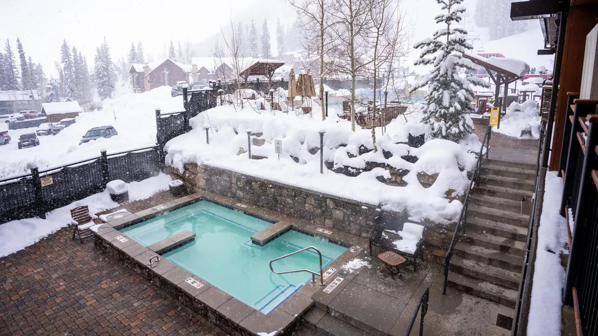 This image depicts an inviting outdoor hot tub surrounded by snow, trees, and an outdoor seating area. The setting conveys the unique experience of relaxing in warm water while taking in the tranquil winter scenery, with the contrast of steam rising from the tub against the snowy environment.