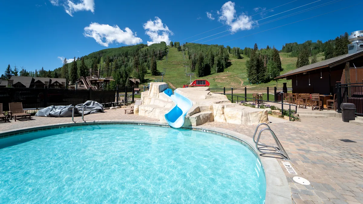 An inviting swimming pool scene with a water slide, lounge chairs, and the majestic mountains in the distance. The sunlit area suggests relaxation, social gatherings, and fun after an adventurous day.