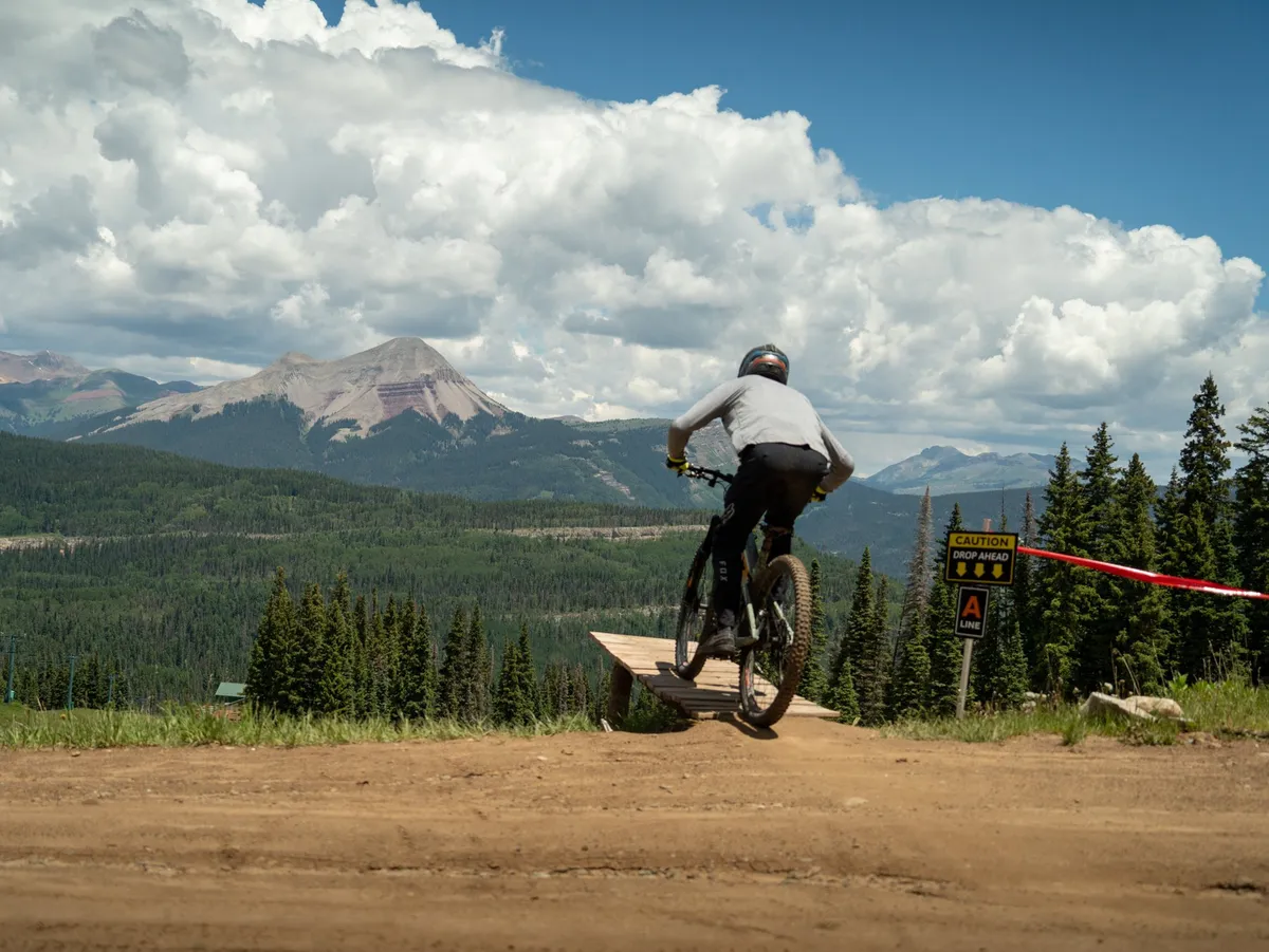 An action-focused scene with a mountain biker riding off a wooden ramp against the backdrop of rugged mountain peaks and clear skies. A caution sign and natural terrain suggest the excitement and challenge of summer mountain activities.