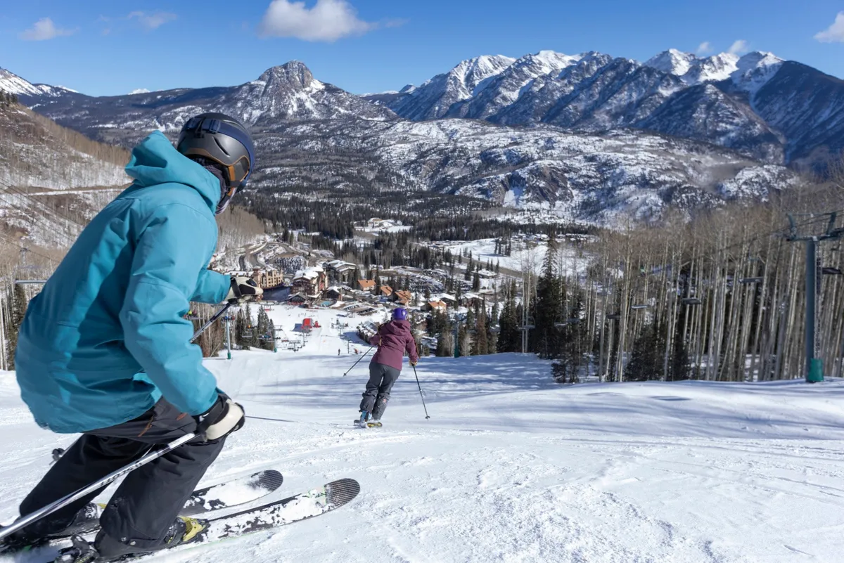 A wide shot of two skiers in vibrant jackets descending a snow-covered slope with a dramatic mountain range and the ski resort visible below. The energy of their movement and the panoramic alpine backdrop capture the thrill of mountain adventures and winter sports.