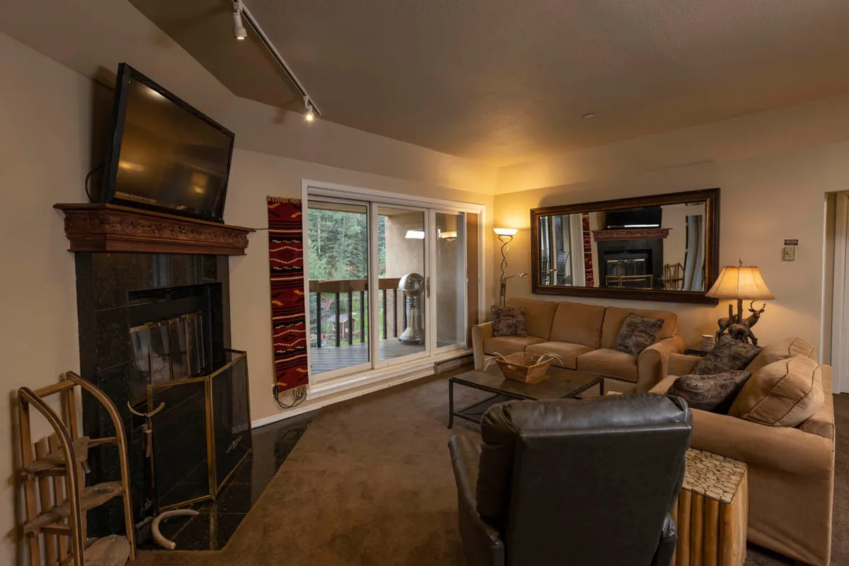 The image showcases a cozy, warmly lit living area featuring a crackling fireplace, plush sofas for lounging, a large mirror reflecting the inviting space, and glass doors leading out to a deck with mountain views. The scene exudes comfort, warmth, and relaxation, perfect for après-ski moments.