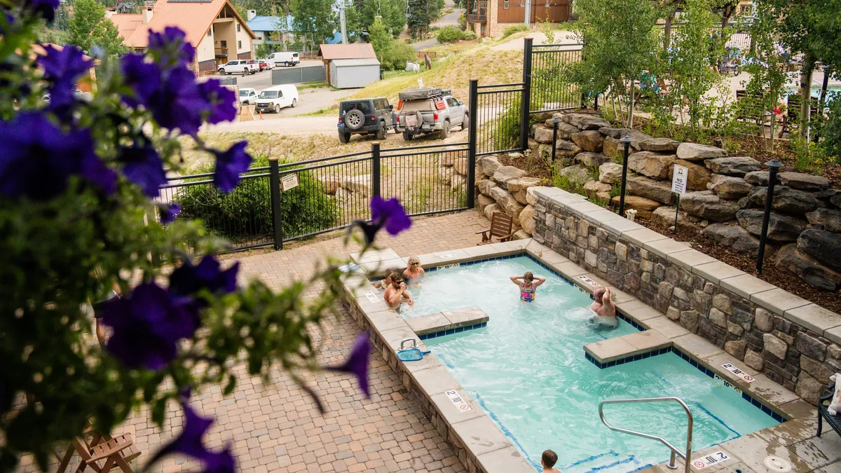 A vibrant exterior shot of the Purgatory Lodge pool and hot tub area, with people enjoying the water, surrounded by stone walls and colorful flowers. The scene suggests relaxation and shared family experiences after a day on the slopes.