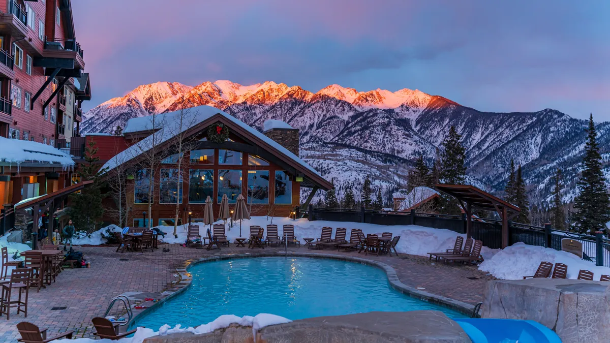 A serene swimming pool sits amidst snow-capped mountains and elegant lodge architecture, with patio furniture set for relaxation. The scene combines the allure of refreshing swims with the visual appeal of a high-end mountain resort.