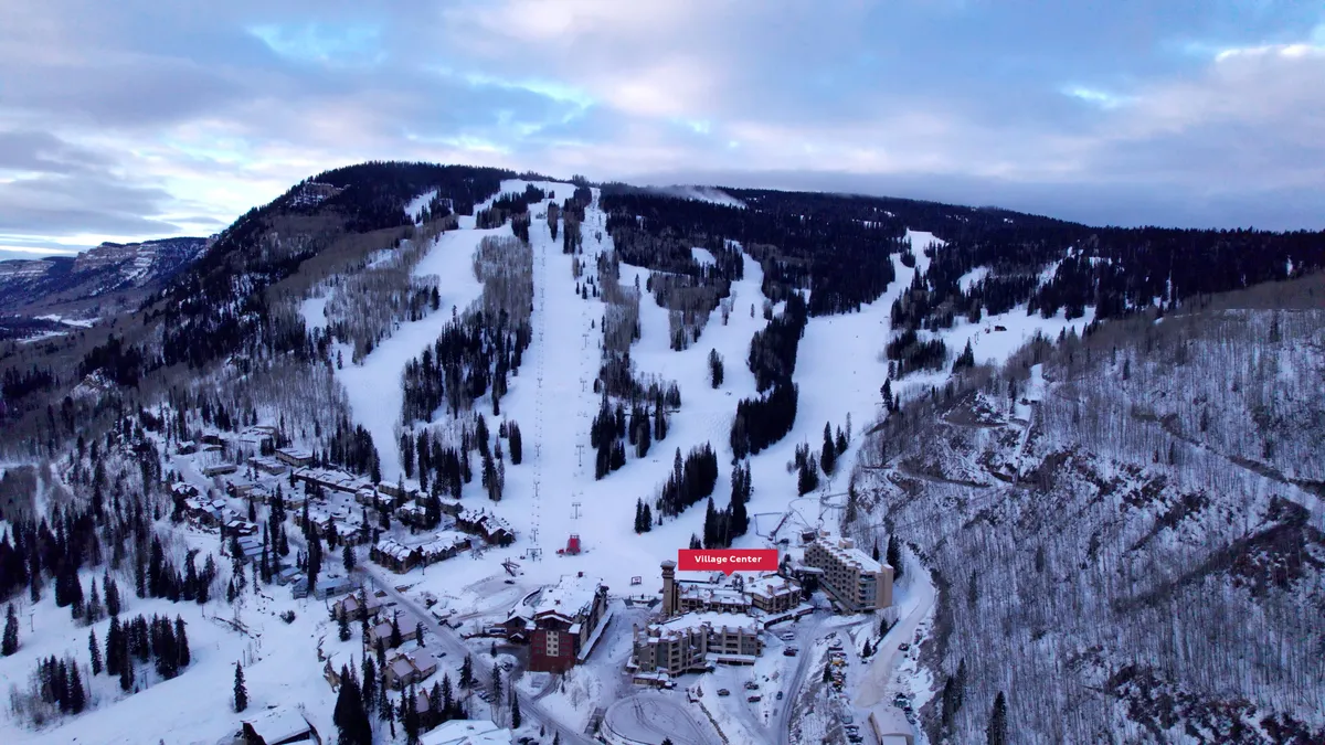 This image captures the ski slopes right outside the Village Center, framed by snow-covered trees and surrounding mountains. The Village Center is visible, emphasizing its proximity to the action. The overall effect is one of adventure and easy access to winter sports.