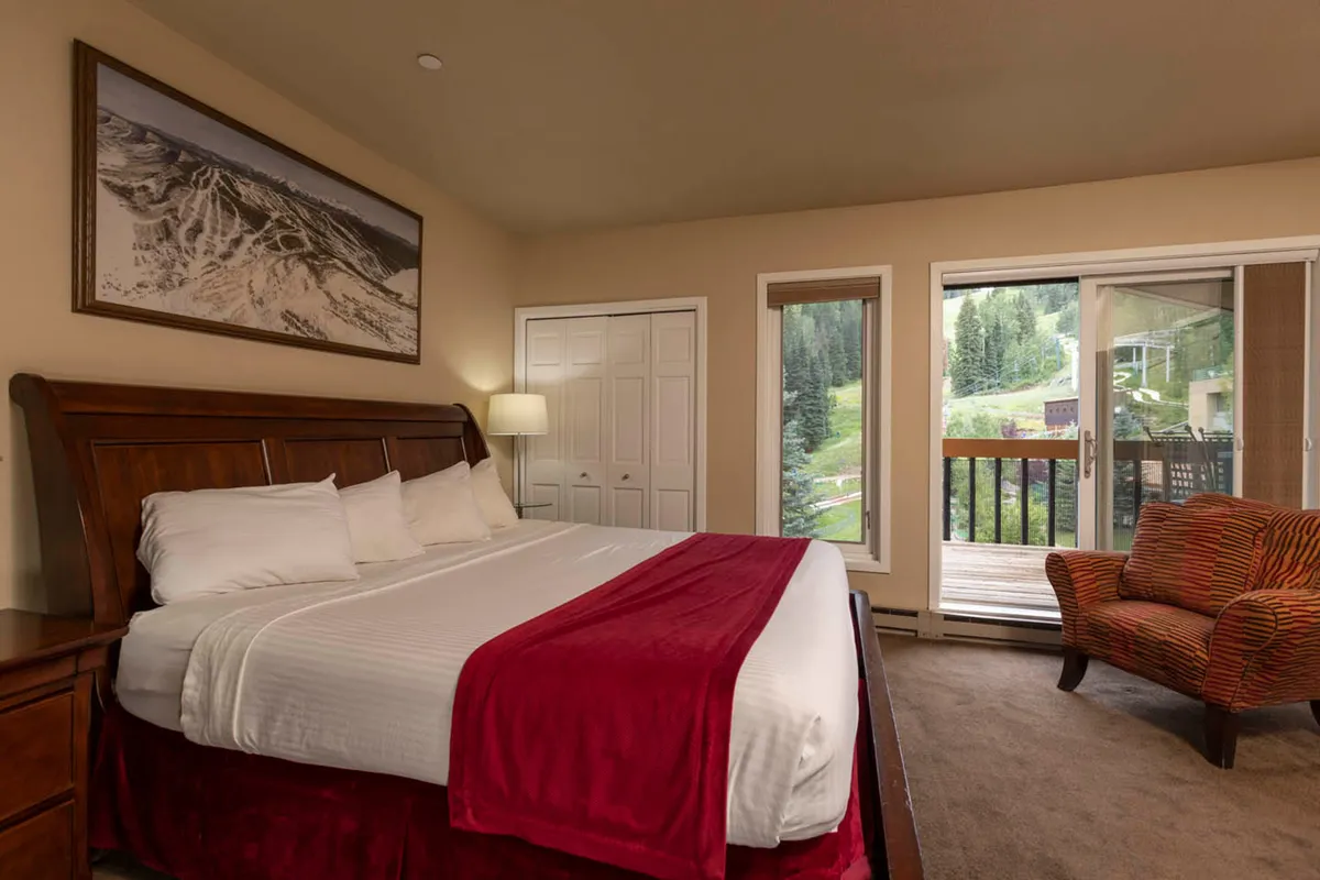This image features a king-sized bed with a red accent blanket, a striped armchair, and large windows overlooking a scenic mountain view. The mountain landscape artwork above the bed and the overall decor convey a tranquil, restful atmosphere, ideal for unwinding after outdoor adventures.